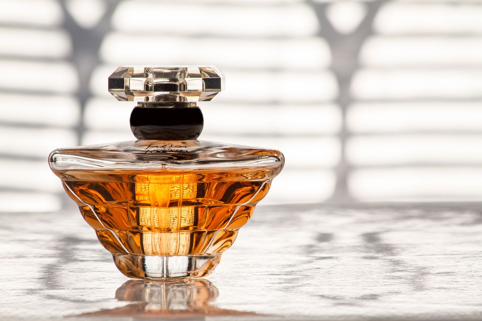 A glass perfume bottle filled with amber fragrance, sunlight falling through window blinds behind it